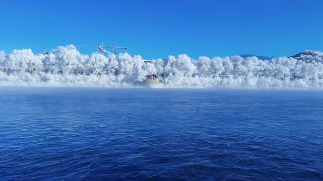 JILIN, CHINA - FEBRUARY 11 2026: A cinematic horizontal shot taken from a moving boat on the Songhua River. The footage captures the breathtaking rime ice covering trees along the riverbank under a cl