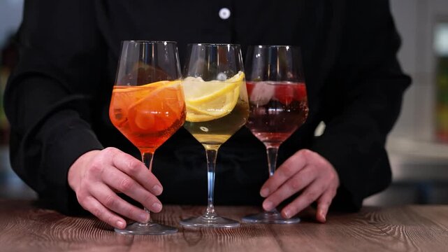 Bartender revealing three colorful citrus cocktails on wooden bar counter in elegant sparkling stem glasses