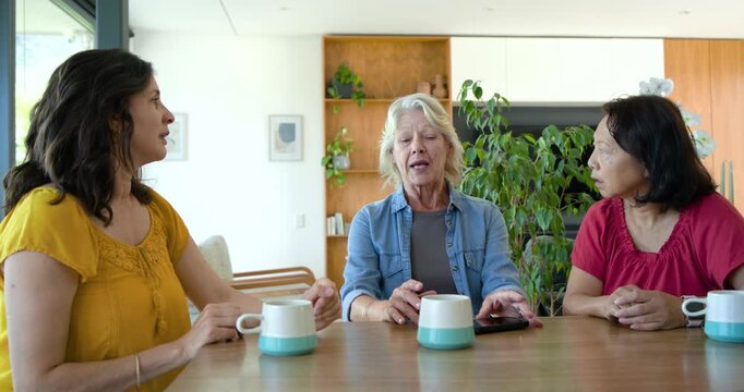 Diverse women including senior gesturing speaker sharing news at table with mugs after phone ping