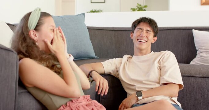 Diverse couple sitting on gray sofa at home, reacting after playful comment, laughing and gesturing