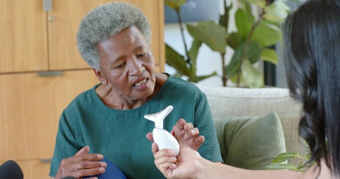 African American caregiver offering device for demo while senior woman testing on cheek on couch