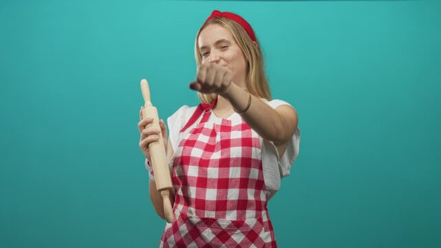 Woman chef in red gingham apron holds a rolling pin and extends a clenched fist toward camera in studio setting; playful.