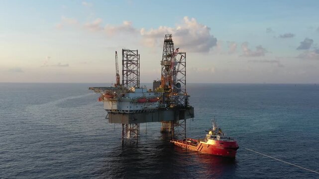 Aerial view from a drone of big jack up offshore drilling oil rig, production platform and supply vessel In the ocean during sunset - Oil and Gas Industry
