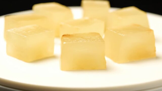 Macro shot of yellow translucent gelatin or jelly cubes neatly arranged on a white serving plate, featuring a bright, high-contrast studio setting.