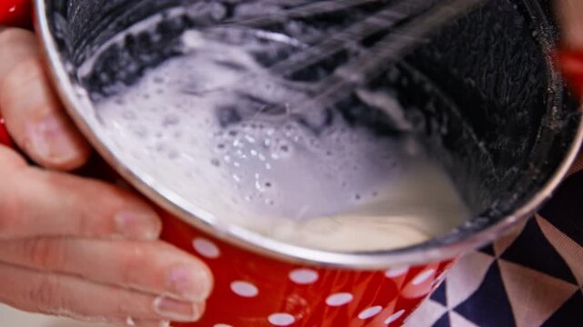 A person holds a red bowl with white polka dots while whisking foamy batter inside, showing a close-up of hands during a food preparation proces