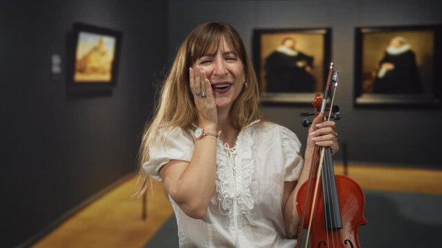 Woman smiling and holding violin with hand on cheek gesture while standing near framed paintings in gallery building; joyful celebration.