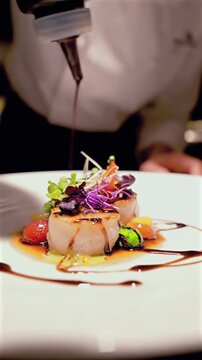 Chef plating scallop with balsamic drizzle, focused female chef in white jacket applies precise sauce lines over seared scallop, microgreens and colorful vegetables arranged, closeup hands