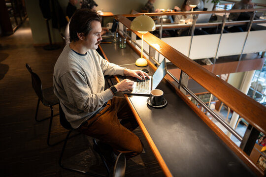 Concentrated businessman working on laptop in trendy co-working space, typing on computer reading data and project information. Focused male freelancer works on remote with computer in cozy cafe.