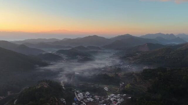 A cinematic horizontal aerial shot captures a tranquil village nestled in a deep valley during sunset. The sky fades from orange to deep blue over distant mountain ridges, creating a peaceful rural at