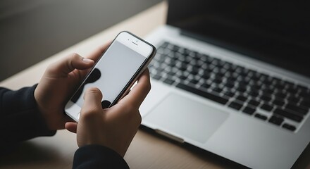 Person using smartphone while a laptop sits nearby on a desk.