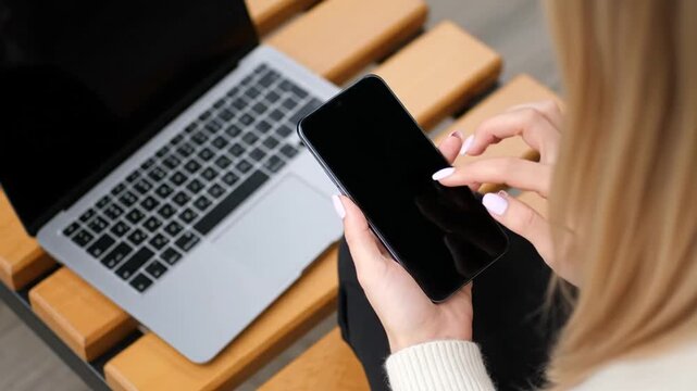 A person with blonde hair using a smartphone. A laptop is next to the phone on a wooden bench