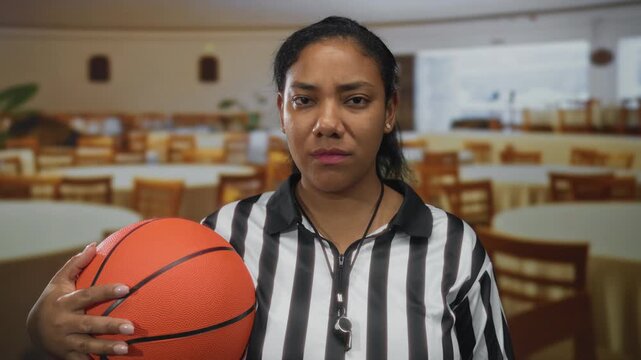 Woman referee wearing striped uniform holds basketball and shows card in building; fairness seriousness.