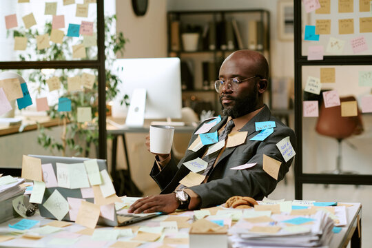 Black man sitting at desk working on laptop surrounded by sticky notes holding coffee cup appearing focused in busy office environment during business day