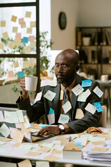 Black man wearing suit sitting at desk covered in sticky notes, holding coffee mug, working on...
