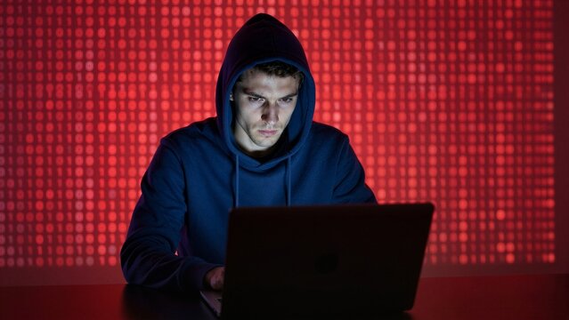 Young Caucasian male in a dark hoodie using a laptop against a red binary code background for advanced cybersecurity threat monitoring and data breach prevention.