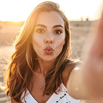 A young woman takes a selfie outdoors. The image is a close-up, revealing her face as she poses with a kissy expression. The background has a sunset glow