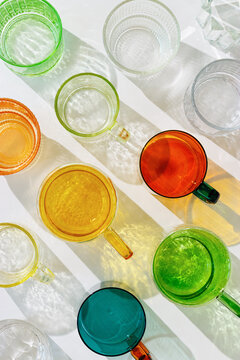Colorful glowing glass cups on white background, casting artistic light reflections and shadows in natural sunlight. Summer drink concept, minimal trend pattern of transparent glasses, caustic