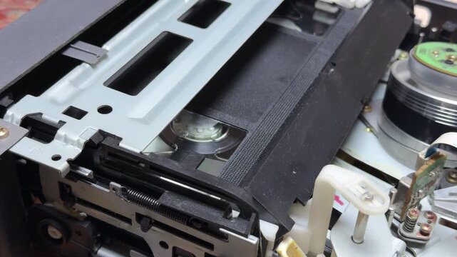 A vintage VCR mechanism pulls a VHS tape into position for playback. The moving gears and metal rollers produce a rhythmic mechanical clatter and a soft, steady whirring sound.