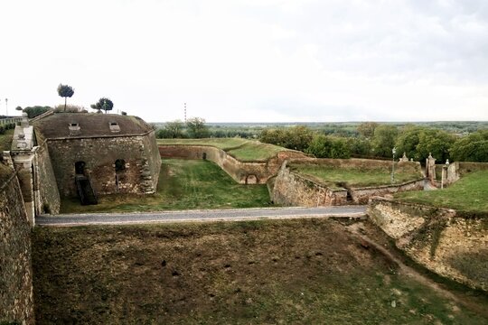 View of the historic Petrovaradin Fortress with stone ramparts, bastions, dry moats and a paved road under a cloudy sky in Novi Sad, Serbia. The European military fortification and defensive structure