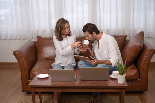 Loving wife feeding coffee to husband while working together in living room. Happy couple sitting on leather sofa with laptops and clipboard. Concept of care, support, and work from home lifestyle.