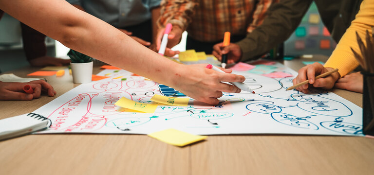 A dynamic brainstorming session showcases diverse hands actively contributing ideas with colorful markers and sticky notes on large paper in a collaborative office environment. SACTR