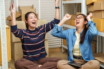 Obraz premium asian women partners raising arms and cheering after successful ecommerce order management celebrating online business achievement inside home warehouse with stacked parcel boxes