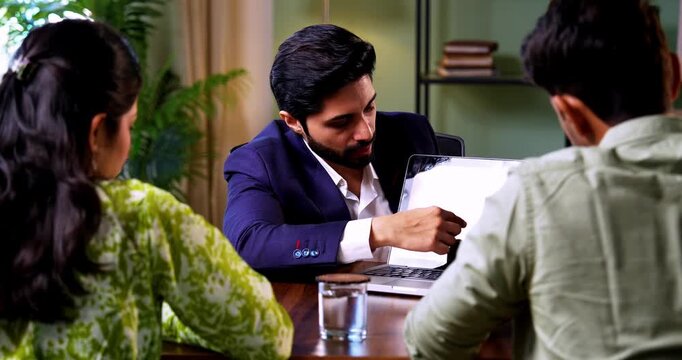 Indian couple visiting bank officer showing laptop screen sitting across table during financial consultation discussing investment and real estate plans in modern corporate office interior