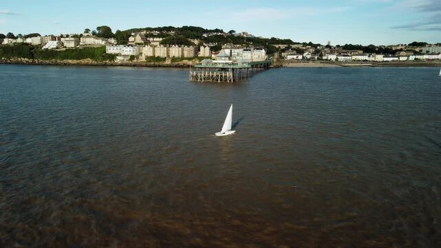 A drone shot towards Clevedon Pier with sail boats in the foreground and the coastline in the background. The shot then does a smooth 360 around the end of the pier