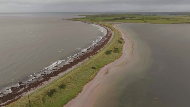 A highangle drone shot capturing the narrow sandspit of Fenit Island in County Kerry, Ireland, featuring crashing waves and green pastures under a moody sky.