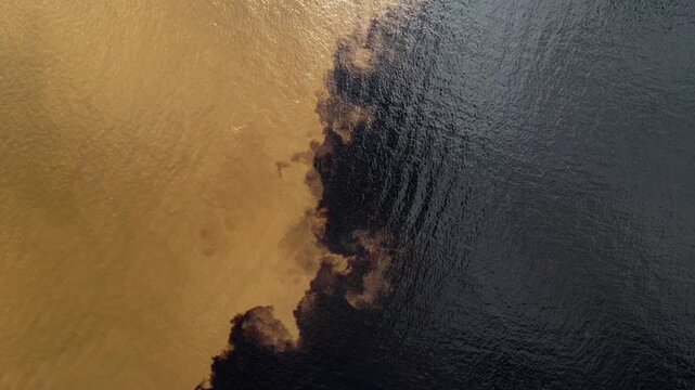 Top-down aerial recede over striking color limits of Black and Amazon river. The murky water resembles liquid gold against the deep blackwater under a cloudy sky.