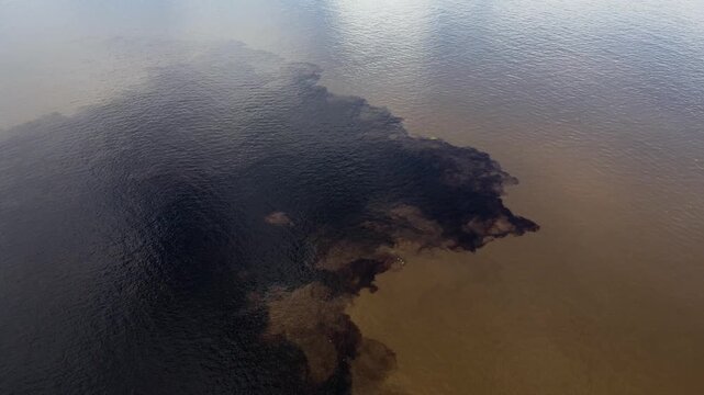 High-angle aerial orbit showing the dramatic convergence of the black Rio Negro and murky Solim&otilde;es. A blue tourist boat traverses the colored patches during bright morning.