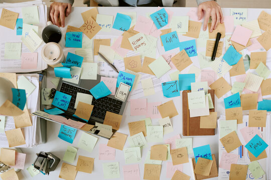Caucasian young adult woman working at cluttered office desk covered with colorful sticky notes, using laptop and reviewing documents, hands visible organizing business tasks