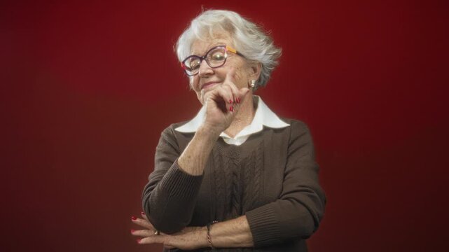 Senior woman points index finger near cheek while crossing arms, wearing glasses and brown sweater against red studio wall; confidence wisdom.