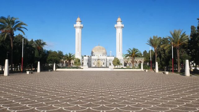 Habib Bourguiba Mausoleum in Monastir, Tunisia featuring golden dome and twin minarets, symmetrical front view across patterned courtyard under clear blue sky with palm trees