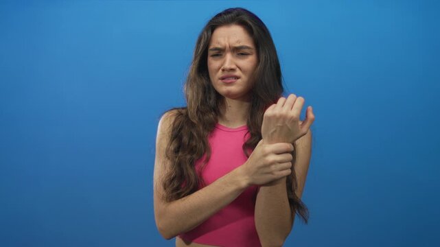 Teenage hispanic woman holds wrist with furrowed brow showing pain gesture in blue studio; distress.