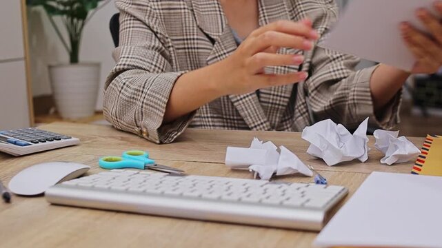 Close-up of businesswomen hands crumpling white paper at office desk with keyboard and scissors

