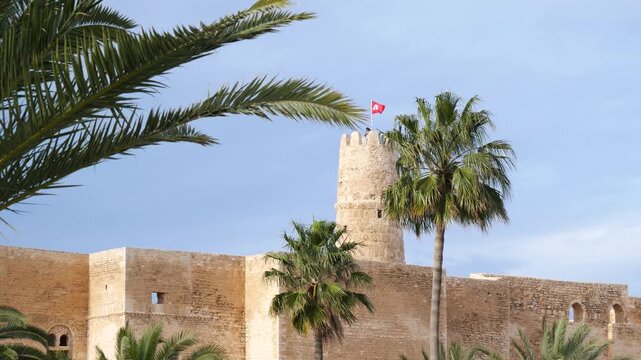 Ribat of Monastir in Tunisia with stone fortress walls and watchtower flying national flag, framed by palm trees under dramatic sky, highlighting historic Islamic architecture