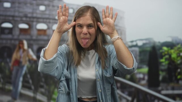 Young blonde woman in denim jacket sticks tongue out joyfully in front of ancient coliseum building; playfulness.