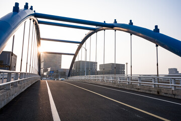 Naklejka premium Blue arched bridge at sunrise with empty urban road and modern cityscape in background