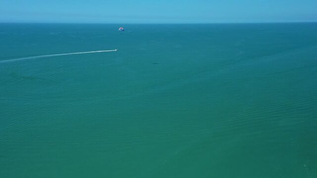 Boat pulls a parachute-like parasail over calm blue ocean water creating wake trails, illustrating high-adrenaline water sport activity under clear skies and open sea conditions.