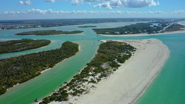 Estuary with green waters and a wide sandy barrier island surrounded by small boats anchored along the shoreline under partly cloudy skies near Florida coast.