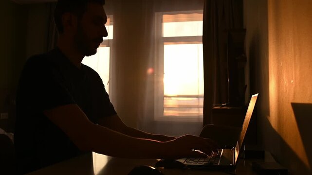 Young freelancer working on a portable computer in a domestic environment, typing on the keyboard as the warm golden light of the setting sun streams through the window behind him