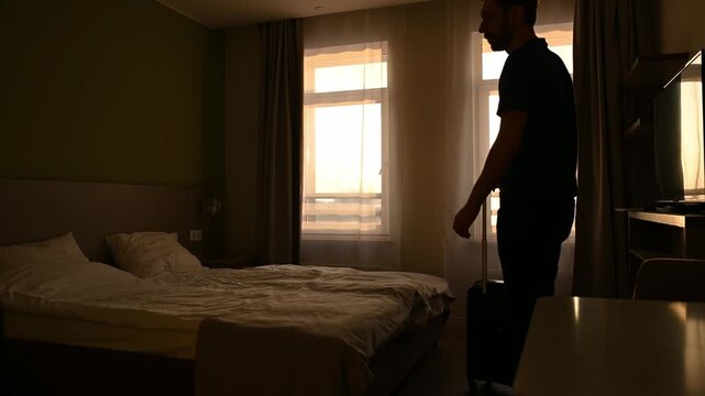 Young man with a suitcase checking into his hotel room after a long trip, silhouetted by the bright light from the window, representing the beginning of a business travel or vacation
