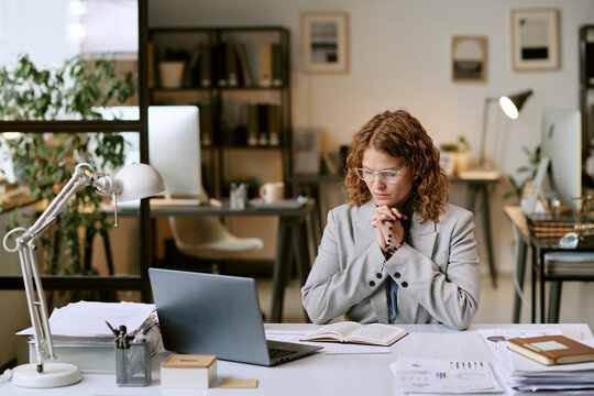 Caucasian woman sitting at desk praying while working on business project, reading open prayer book and concentrating in modern office environment with laptop and documents visible