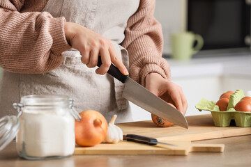 Obraz premium Woman in apron slicing onion on wooden cutting board with chef knife, eggs, garlic and flour jar in home kitchen, cozy domestic food preparation close up