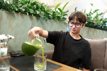 Young man with glasses pouring green smoothie into glass at cafe