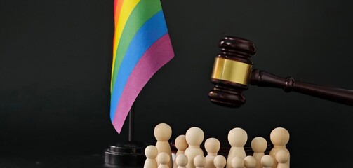 A rainbow flag symbolizing LGBTQ pride stands beside a gavel and wooden figures representing unity in the fight © Nadzeya
