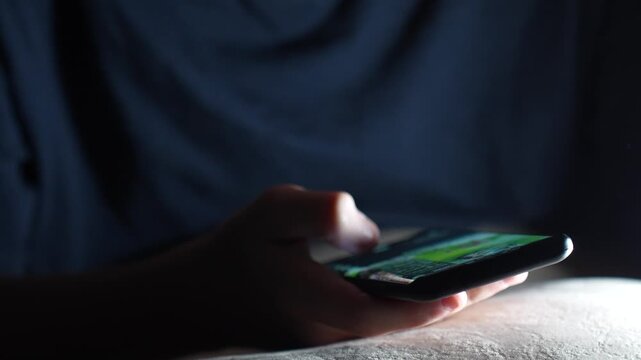 Close-up of Hand Using Smartphone in Dark Room. A low light shot of a person scrolling on a bright smartphone screen at night. Concept for social media addiction, insomnia, and digital communication