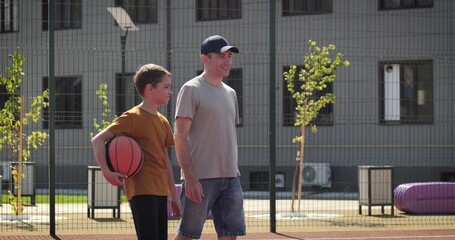 Father accompanies son on outdoor basketball court in city area. Father guides child before sport activity. Warm daylight creates positive atmosphere. Father shows mentoring, care and active lifestyle