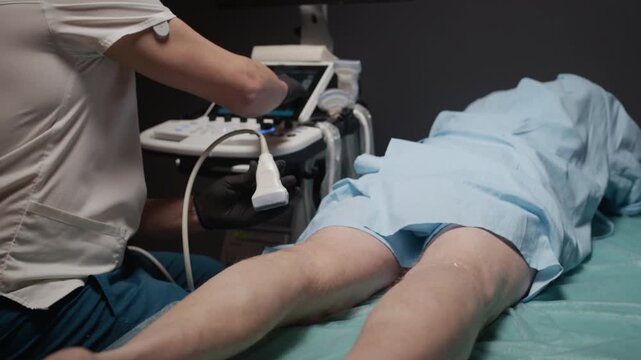 Cropped shot of gloved hands of sonographer evaluating blood flow of legs of female patient lying on examination table, operating ultrasound transducer to capture images displayed on monitor